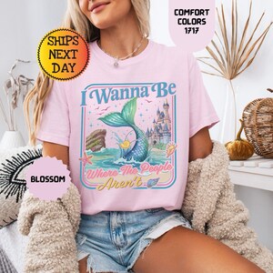 May include: A light pink t-shirt featuring a graphic print of a mermaid tail emerging from the ocean, a castle, and the text I Wanna Be Where The People Aren't. The design includes starfish, seashells, and birds in a rectangular frame.