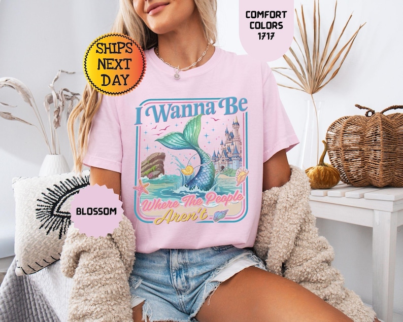 May include: A light pink t-shirt featuring a graphic print of a mermaid tail emerging from the ocean, a castle, and the text I Wanna Be Where The People Aren't. The design includes starfish, seashells, and birds in a rectangular frame.