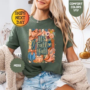 May include: A moss green t-shirt featuring a graphic print with the text It's Fall Y'all, a brown cowboy hat, a blue pumpkin, an orange pumpkin, floral accents, and a pair of brown cowboy boots with floral patterns on the front of the shirt.