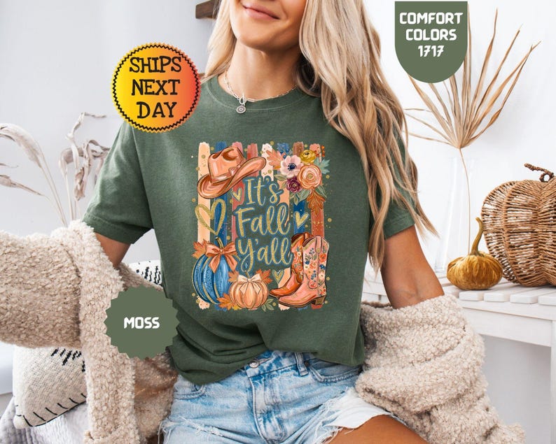 May include: A moss green t-shirt featuring a graphic print with the text It's Fall Y'all, a brown cowboy hat, a blue pumpkin, an orange pumpkin, floral accents, and a pair of brown cowboy boots with floral patterns on the front of the shirt.