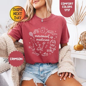 May include: Crimson-colored t-shirt with the text "movement is medicine" and fitness-related graphics in white. The shirt features a crew neck and short sleeves. The image also includes the words "Comfort Colors" and "Ships Next Day".