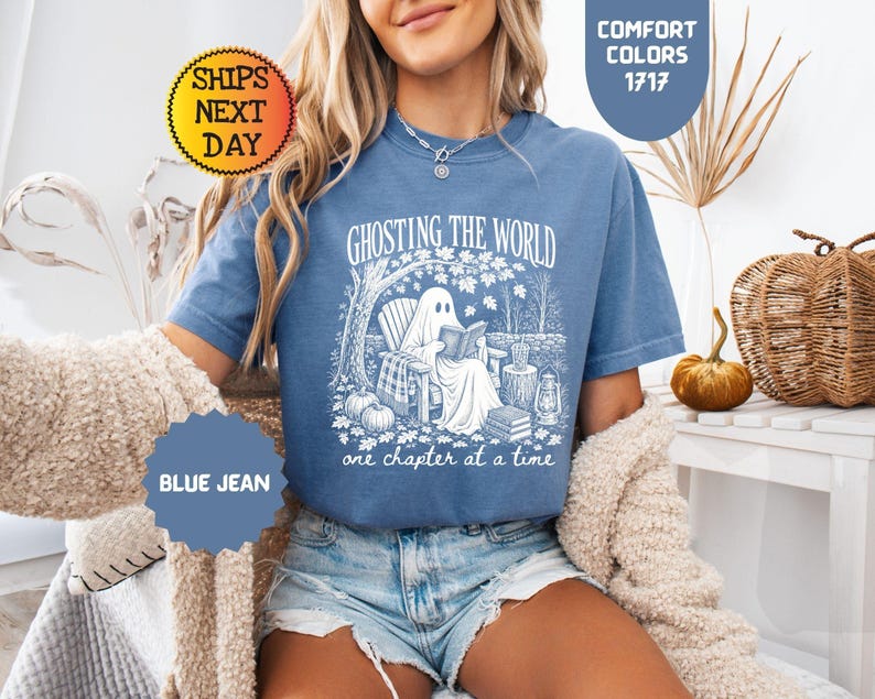 May include: A blue jean colored t-shirt featuring a white graphic print of a ghost sitting in a wooden chair reading a book under a tree. The text reads GHOSTING THE WORLD one chapter at a time. The shirt has a crew neckline and short sleeves.