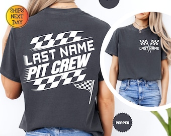 Puede incluir: Una camiseta gris carbón de manga corta con gráficos serigrafiados en blanco. La parte trasera muestra motivos de banderas a cuadros con el texto LAST NAME PIT CREW. El pecho izquierdo presenta un gráfico más pequeño de dos banderas y el texto LAST NAME.