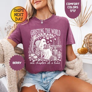 May include: A berry-colored short-sleeve t-shirt featuring a white graphic print of a ghost sitting in a chair reading a book under a tree. The text reads GHOSTING THE WORLD one chapter at a time. The shirt is worn with light blue denim shorts.