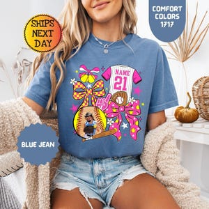 May include: A blue jean colored t-shirt featuring a graphic print of a softball jersey with the text NAME 21, a softball, a baseball glove, a wooden bat, and large pink and yellow checkered bows. The shirt is labeled Comfort Colors 1717.