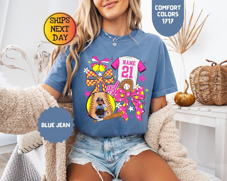 May include: A blue jean colored t-shirt featuring a graphic print of a softball jersey with the text NAME 21, a softball, a baseball glove, a wooden bat, and large pink and yellow checkered bows. The shirt is labeled Comfort Colors 1717.
