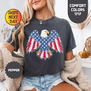 May include: Dark gray t-shirt featuring an American flag eagle design. The eagle's wings display red and white stripes and blue stars. The shirt has the text "Comfort Colors 1717" and "Pepper".