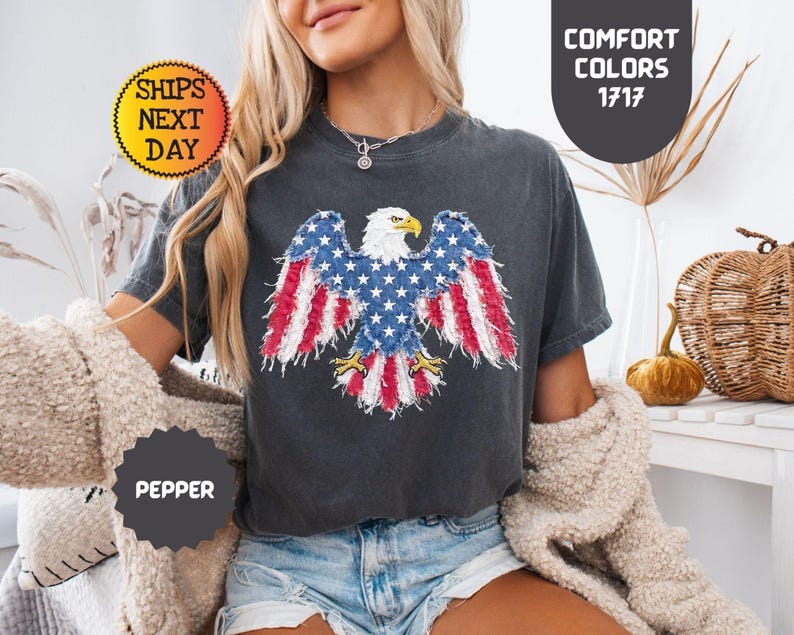 May include: Dark gray t-shirt featuring an American flag eagle design. The eagle's wings display red and white stripes and blue stars. The shirt has the text "Comfort Colors 1717" and "Pepper".