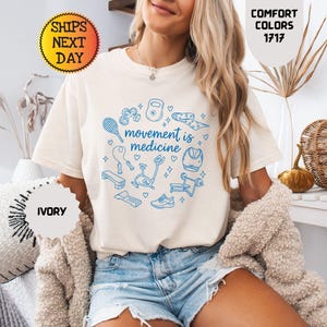 May include: Ivory-colored t-shirt with the text "movement is medicine" surrounded by blue fitness-related illustrations. The shirt is part of the Comfort Colors 1717 collection. The image also includes the text "Ships Next Day" and "Ivory".