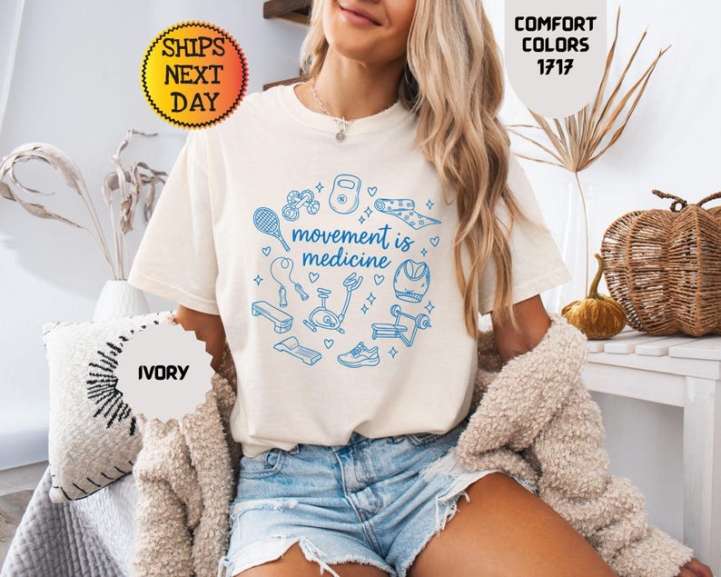 May include: Ivory-colored t-shirt with the text "movement is medicine" surrounded by blue fitness-related illustrations. The shirt is part of the Comfort Colors 1717 collection. The image also includes the text "Ships Next Day" and "Ivory".