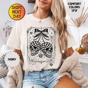 以下が含まれることがあります： アイボリーの半袖Tシャツ。大きなリボンから吊り下げられた2つのチェッカー柄の頭蓋骨の黒いグラフィックプリントが特徴です。デザインには小さなハートのアクセント、星、装飾的なフィリグリーの縁取りが含まれています。Comfort Colors 1717というテキストが表示されています。