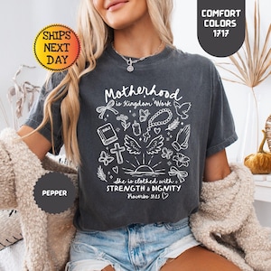 May include: Dark gray t-shirt with the text "Motherhood is Kingdom Work" and the quote "She is clothed with strength & dignity Proverbs 31:25" in white. The shirt has a graphic of a bible, cross, and other religious symbols. The shirt is a comfort color.