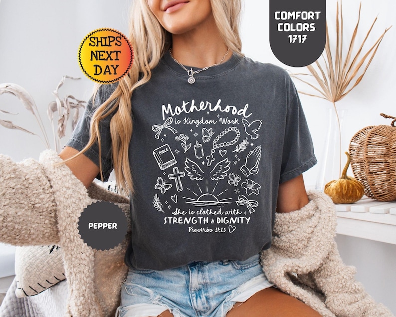 May include: Dark gray t-shirt with the text "Motherhood is Kingdom Work" and the quote "She is clothed with strength & dignity Proverbs 31:25" in white. The shirt has a graphic of a bible, cross, and other religious symbols. The shirt is a comfort color.