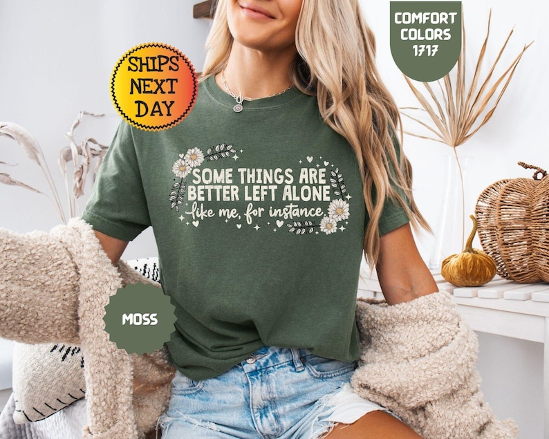 May include: Moss green t-shirt with the text "SOME THINGS ARE BETTER LEFT ALONE like me, for instance." The shirt has a floral design and is part of the Comfort Colors collection. The image also includes a "Ships Next Day" sticker.