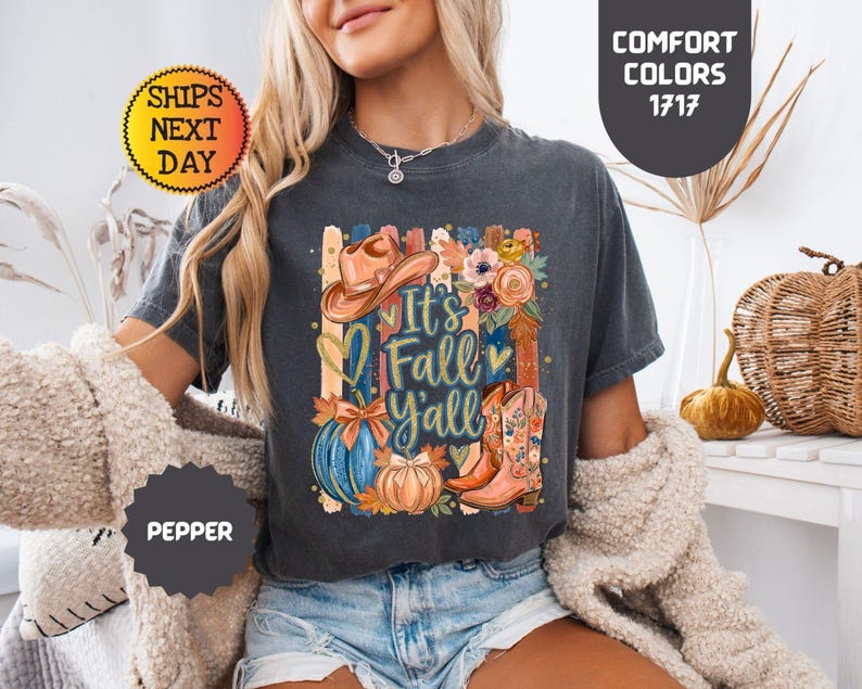 May include: A charcoal gray short-sleeve t-shirt featuring a graphic print with the text It's Fall Y'all, a cowboy hat, a blue pumpkin, an orange pumpkin, a floral arrangement, and a western boot with floral patterns on the side.