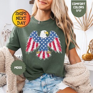 May include: A moss green t-shirt featuring an embroidered American flag eagle design. The eagle's wings display the red and white stripes and blue stars of the flag. The shirt also has the text "Comfort Colors 1717" and "Ships Next Day".