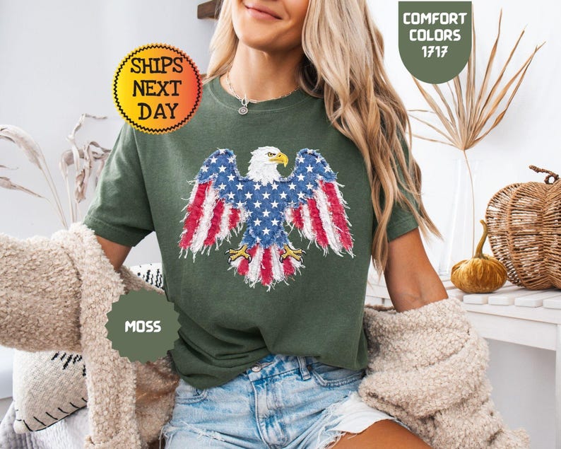 May include: A moss green t-shirt featuring an embroidered American flag eagle design. The eagle's wings display the red and white stripes and blue stars of the flag. The shirt also has the text "Comfort Colors 1717" and "Ships Next Day".