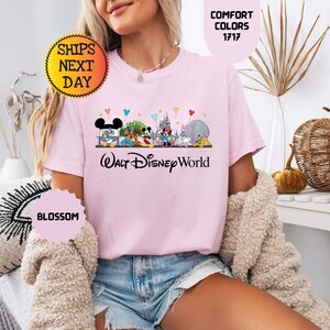 May include: A light pink t-shirt featuring a colorful graphic of Disney characters and the text "Walt Disney World". The shirt also has the text "Comfort Colors 1717" and "Blossom".