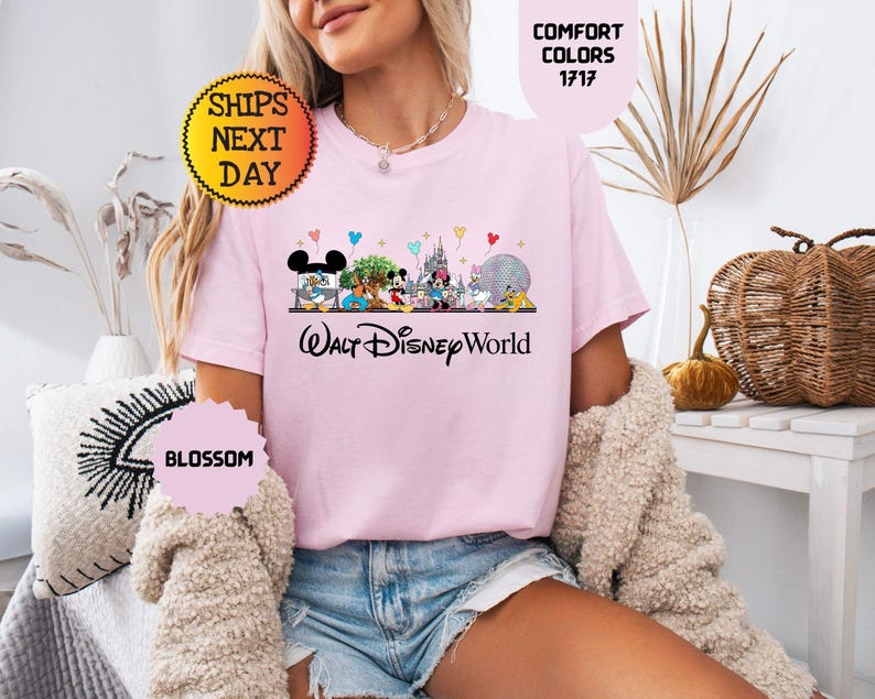 May include: A light pink t-shirt featuring a colorful graphic of Disney characters and the text "Walt Disney World". The shirt also has the text "Comfort Colors 1717" and "Blossom".