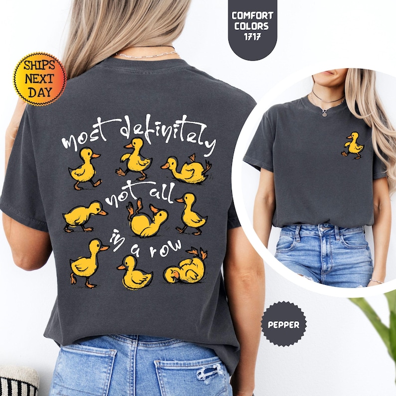 May include: Dark gray t-shirt with the phrase "most definitely not all in a row" and cartoon ducklings. A small duckling graphic is on the front. The shirt is a Comfort Colors 1717, and the color is Pepper.