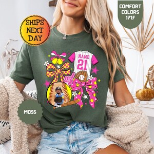 May include: A moss green t-shirt featuring a graphic print of a softball jersey with the text NAME 21, a baseball glove, a softball, a wooden bat, a foam finger, and two large pink and yellow checkered bows with small softball icons.
