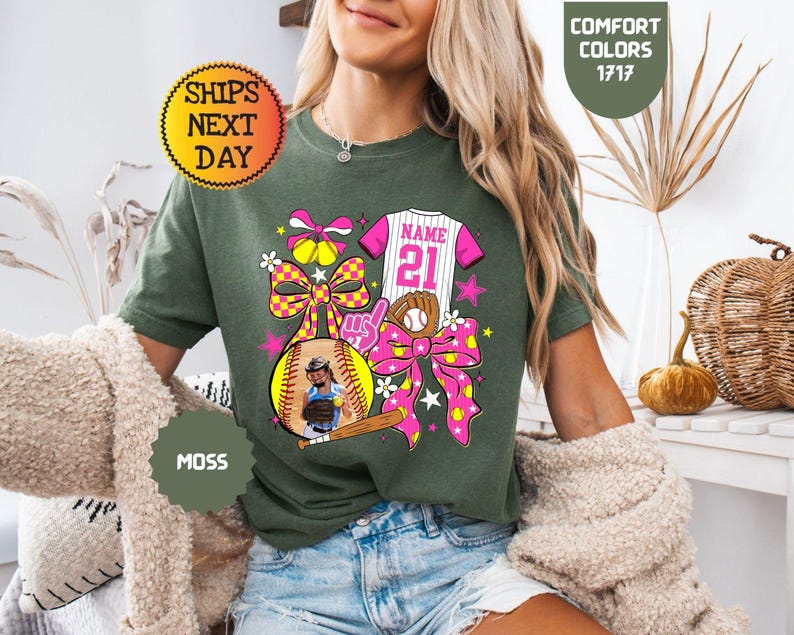 May include: A moss green t-shirt featuring a graphic print of a softball jersey with the text NAME 21, a baseball glove, a softball, a wooden bat, a foam finger, and two large pink and yellow checkered bows with small softball icons.