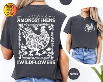 Könnte beinhalten: Ein dunkelgraues T-Shirt mit einer weißen Grafik einer Henne, umgeben von Blumen und dem Text "you will find me amongst the hens and wildflowers".