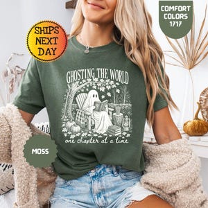 May include: A moss green short-sleeve t-shirt featuring a white graphic print of a ghost sitting in a chair reading a book under a tree. The text reads GHOSTING THE WORLD one chapter at a time. The shirt is labeled Comfort Colors 1717.