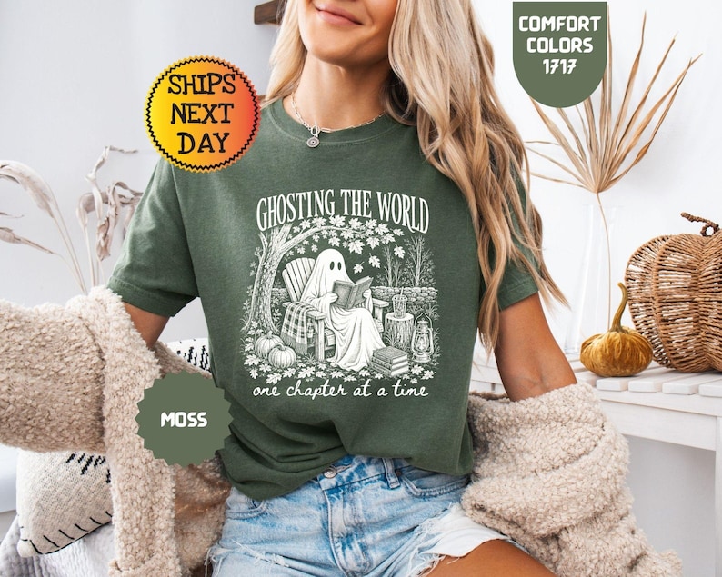 May include: A moss green short-sleeve t-shirt featuring a white graphic print of a ghost sitting in a chair reading a book under a tree. The text reads GHOSTING THE WORLD one chapter at a time. The shirt is labeled Comfort Colors 1717.