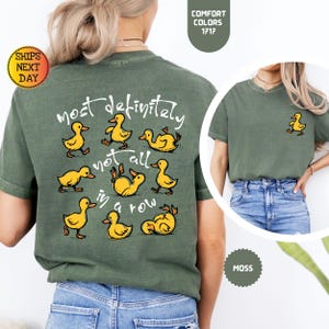 May include: A moss green t-shirt with a playful design featuring yellow cartoon ducks. The back of the shirt displays the phrase "most definitely not all in a row" with duck illustrations. A smaller duck graphic is on the front.