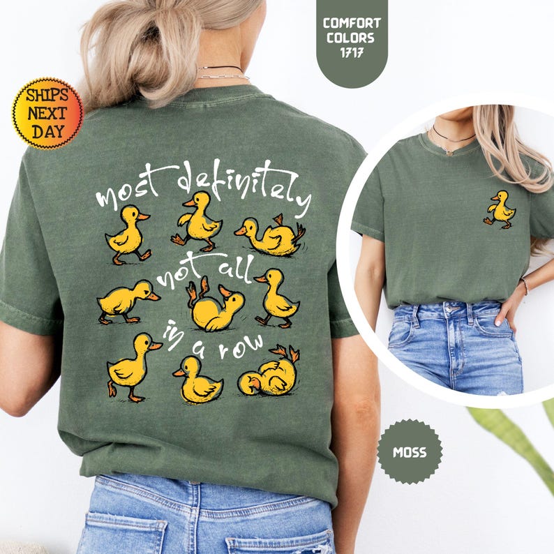 May include: A moss green t-shirt with a playful design featuring yellow cartoon ducks. The back of the shirt displays the phrase "most definitely not all in a row" with duck illustrations. A smaller duck graphic is on the front.