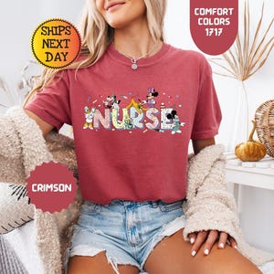 May include: Crimson-colored t-shirt with the word "NURSE" in colorful letters, featuring cartoon characters. The shirt has short sleeves and is paired with denim shorts. The image also includes text that says "Comfort Colors 1717" and "Ships Next Day".