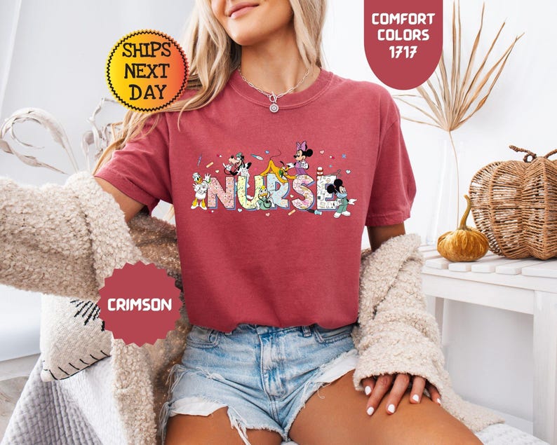 May include: Crimson-colored t-shirt with the word "NURSE" in colorful letters, featuring cartoon characters. The shirt has short sleeves and is paired with denim shorts. The image also includes text that says "Comfort Colors 1717" and "Ships Next Day".