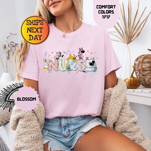May include: A light pink t-shirt with the word "NURSE" in colorful letters, featuring cartoon characters. The shirt has short sleeves and a crew neck. The text "COMFORT COLORS 1717" is visible. The shirt is worn by a person.
