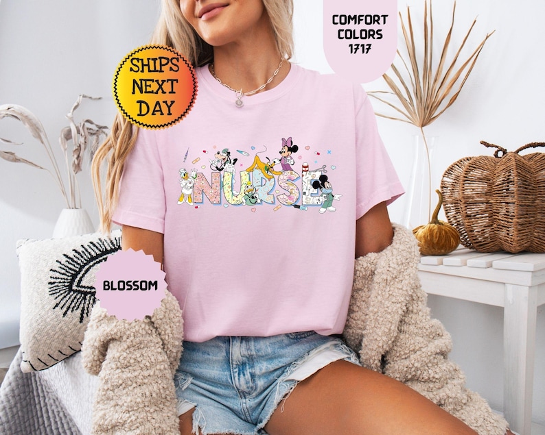 May include: A light pink t-shirt with the word "NURSE" in colorful letters, featuring cartoon characters. The shirt has short sleeves and a crew neck. The text "COMFORT COLORS 1717" is visible. The shirt is worn by a person.