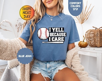 Può includere: Una t-shirt a maniche corte color blu jeans con una grafica raffigurante una palla da baseball e una mazza da baseball. Il testo sulla maglietta recita I YELL BECAUSE I CARE in lettere stampatello nere. Presenta un girocollo.