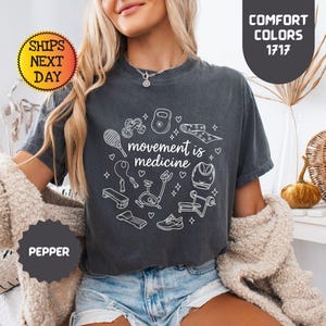 May include: Dark gray t-shirt with the phrase "movement is medicine" surrounded by white line drawings of exercise equipment. The shirt has a crew neck and short sleeves. The text "Comfort Colors 1717" is also visible.
