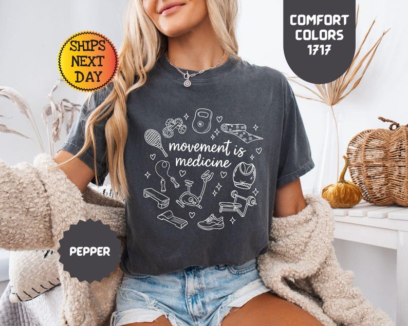 May include: Dark gray t-shirt with the phrase "movement is medicine" surrounded by white line drawings of exercise equipment. The shirt has a crew neck and short sleeves. The text "Comfort Colors 1717" is also visible.