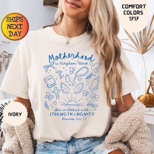May include: Ivory-colored t-shirt with blue graphic text and illustrations. The text reads "Motherhood is Kingdom Work" with additional text from Proverbs 31:25. The shirt also features a cross, wings, and other religious symbols. The shirt is labeled "Comfort Colors 1717".