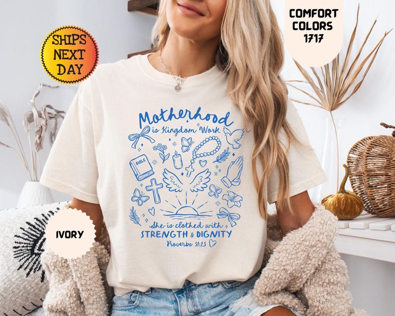 May include: Ivory-colored t-shirt with blue graphic text and illustrations. The text reads "Motherhood is Kingdom Work" with additional text from Proverbs 31:25. The shirt also features a cross, wings, and other religious symbols. The shirt is labeled "Comfort Colors 1717".