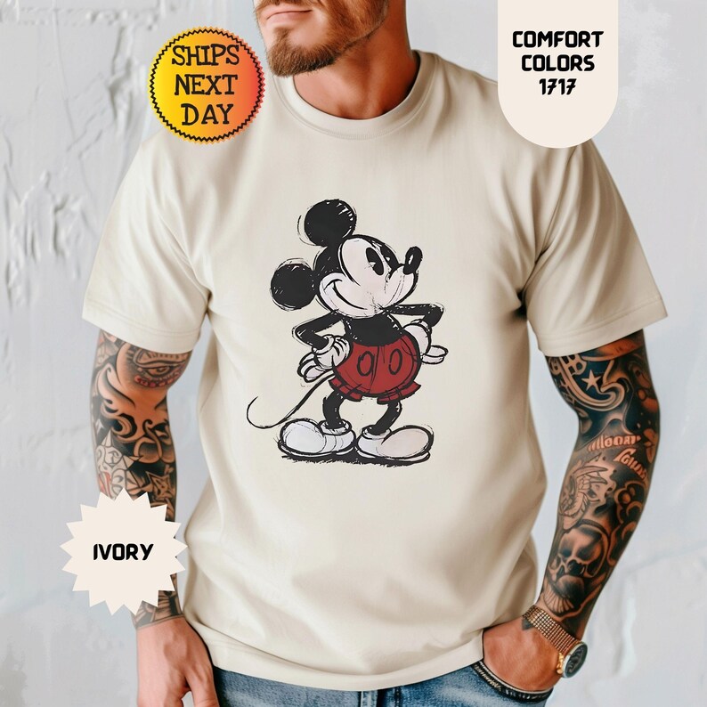 May include: Ivory-colored t-shirt featuring a sketch-style Mickey Mouse graphic in black, red, and white. The shirt has the text "Comfort Colors 1717" and "Ships Next Day".