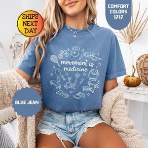 May include: Blue jean colored t-shirt with the text "movement is medicine" surrounded by white line drawings of exercise equipment. The shirt is a comfort color and has a round neck.