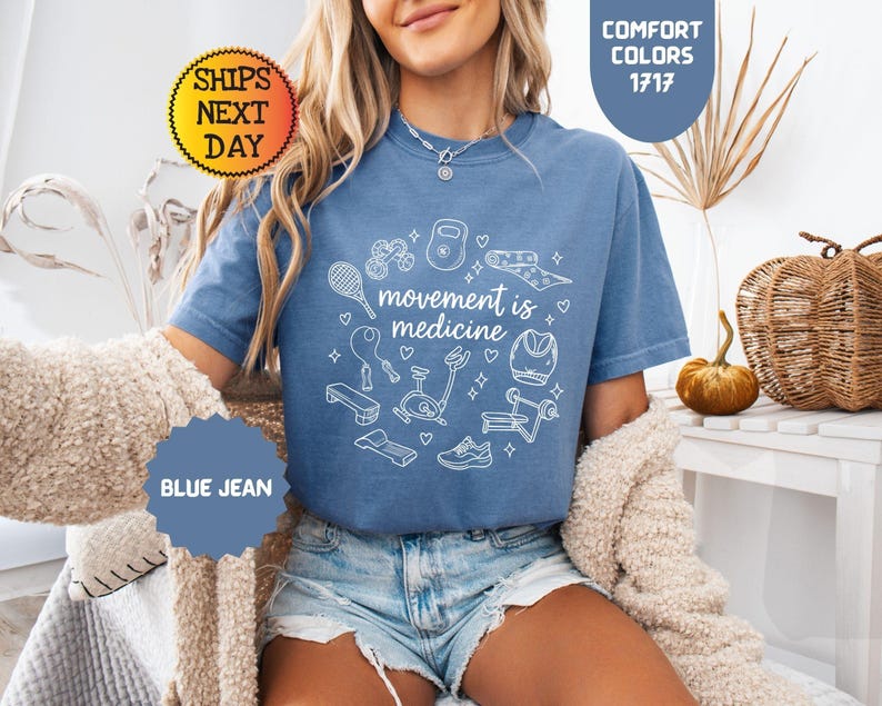 May include: Blue jean colored t-shirt with the text "movement is medicine" surrounded by white line drawings of exercise equipment. The shirt is a comfort color and has a round neck.