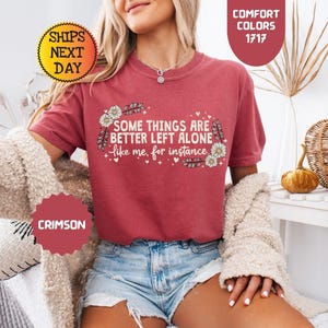 May include: Crimson-colored t-shirt with the text "SOME THINGS ARE BETTER LEFT ALONE like me, for instance" in white. The design includes floral and feather accents. The shirt is part of the Comfort Colors 1717 collection.