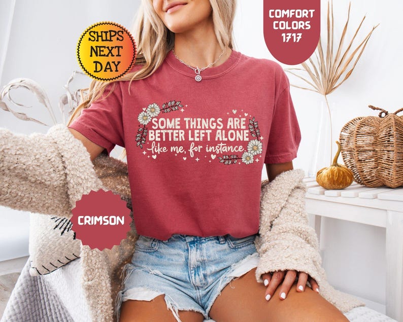 May include: Crimson-colored t-shirt with the text "SOME THINGS ARE BETTER LEFT ALONE like me, for instance" in white. The design includes floral and feather accents. The shirt is part of the Comfort Colors 1717 collection.