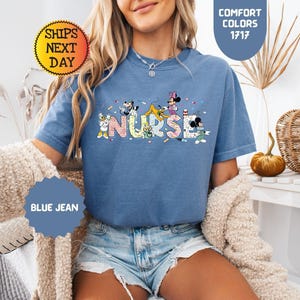 May include: Blue jean colored t-shirt with the word "NURSE" in colorful letters and cartoon characters. The shirt has short sleeves and a crew neck. The text "COMFORT COLORS 1717" is in a blue oval. The text "SHIPS NEXT DAY" is in a yellow circle.