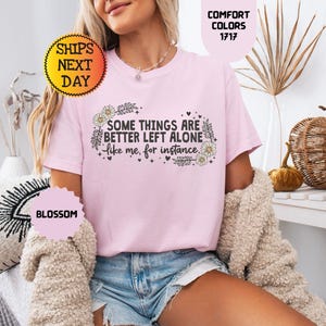 May include: A light pink t-shirt with the text "SOME THINGS ARE BETTER LEFT ALONE like me, for instance" in black. The shirt has a floral design and is part of the "Comfort Colors" collection.