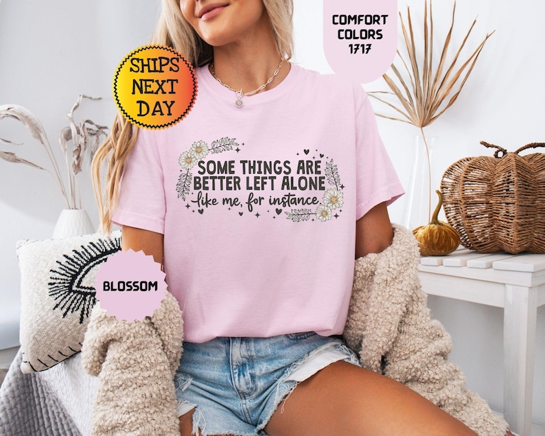May include: A light pink t-shirt with the text "SOME THINGS ARE BETTER LEFT ALONE like me, for instance" in black. The shirt has a floral design and is part of the "Comfort Colors" collection.