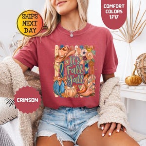 May include: A crimson short-sleeve t-shirt featuring a graphic print with a cowboy hat, a blue pumpkin, a tan pumpkin, and floral-patterned cowboy boots. The text reads It's Fall Y'all, surrounded by vertical stripes and heart shapes.