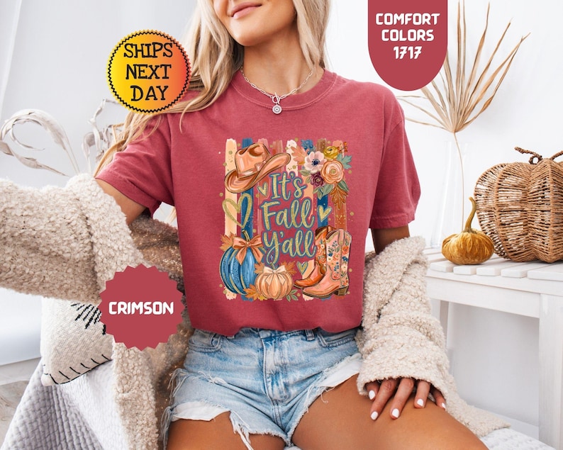 May include: A crimson short-sleeve t-shirt featuring a graphic print with a cowboy hat, a blue pumpkin, a tan pumpkin, and floral-patterned cowboy boots. The text reads It's Fall Y'all, surrounded by vertical stripes and heart shapes.
